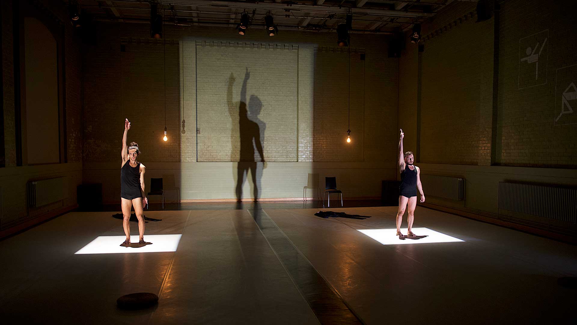 Zwei Tänzer stehen jeweils auf beleuchteten Flächen in einem Bühnenraum und heben einen Arm, während ihre Schatten groß an die Wand projiziert werden.