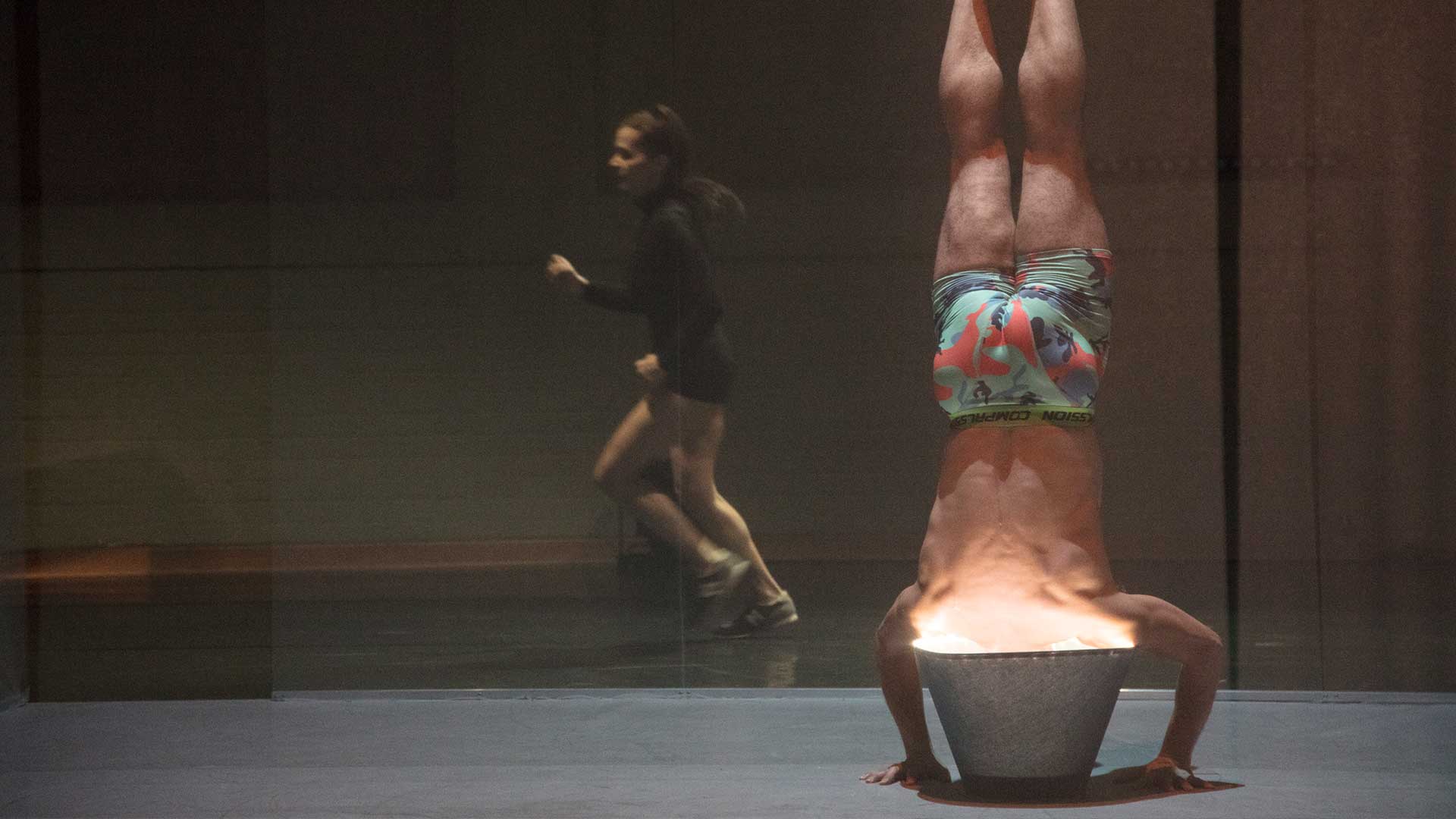 A person holds a headstand with their head immersed in a glowing bowl while another figure moves past blurred in the background.