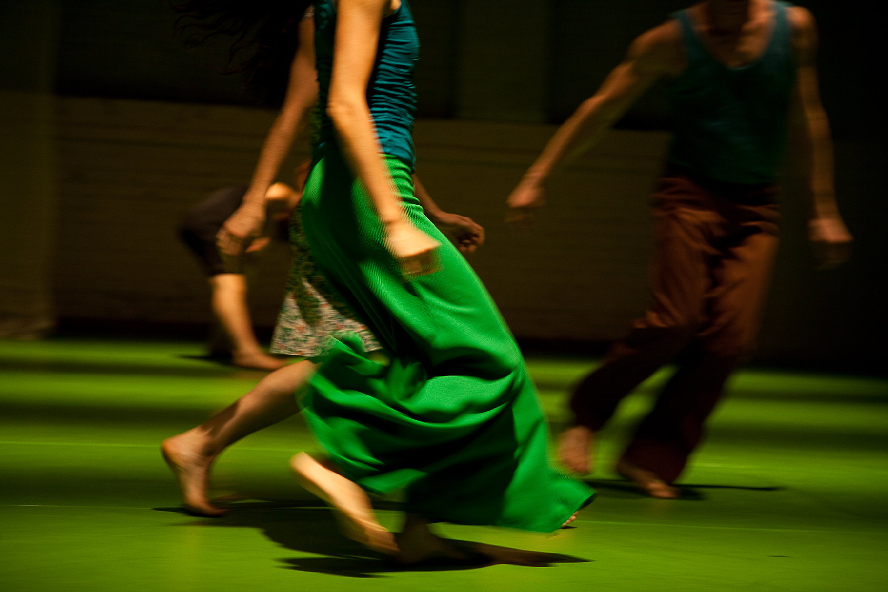 Tänzer:innen in Bewegung auf einer Bühne mit grünem Boden; eine Person im Vordergrund trägt ein langes, fließendes grünes Kleid, während weitere Tänzer:innen unscharf im Hintergrund vorbeilaufen.